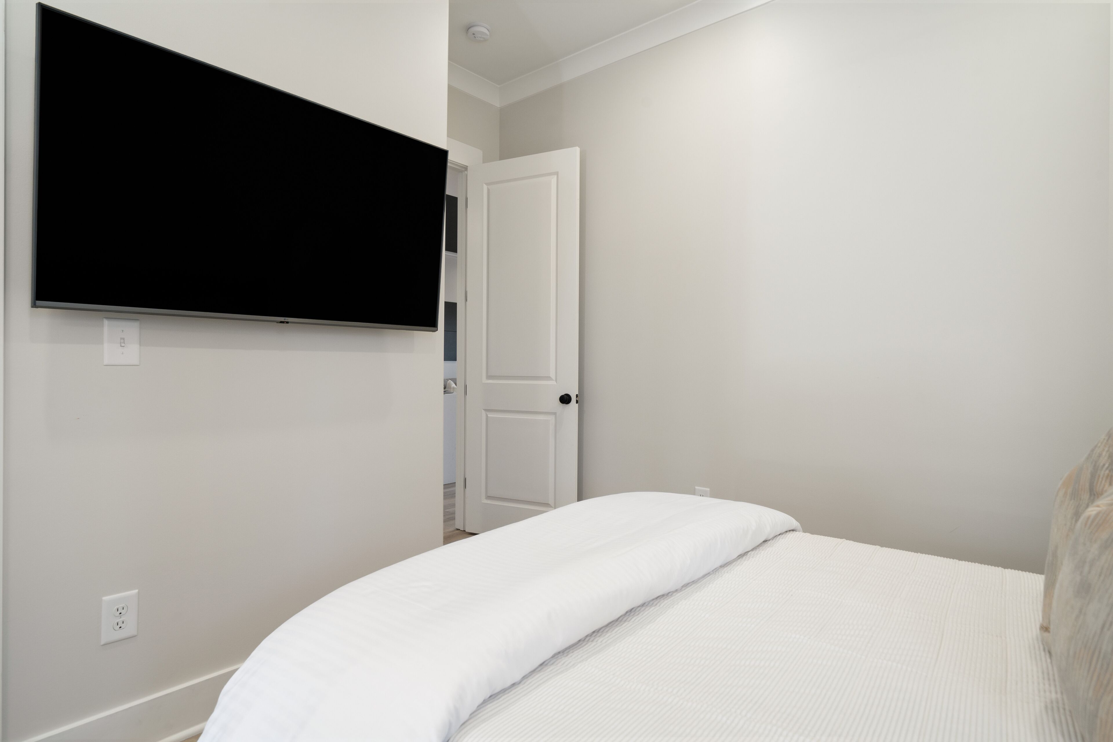 Une chambre à coucher avec un lit, un téléviseur fixé au mur et une porte donnant sur une autre pièce.