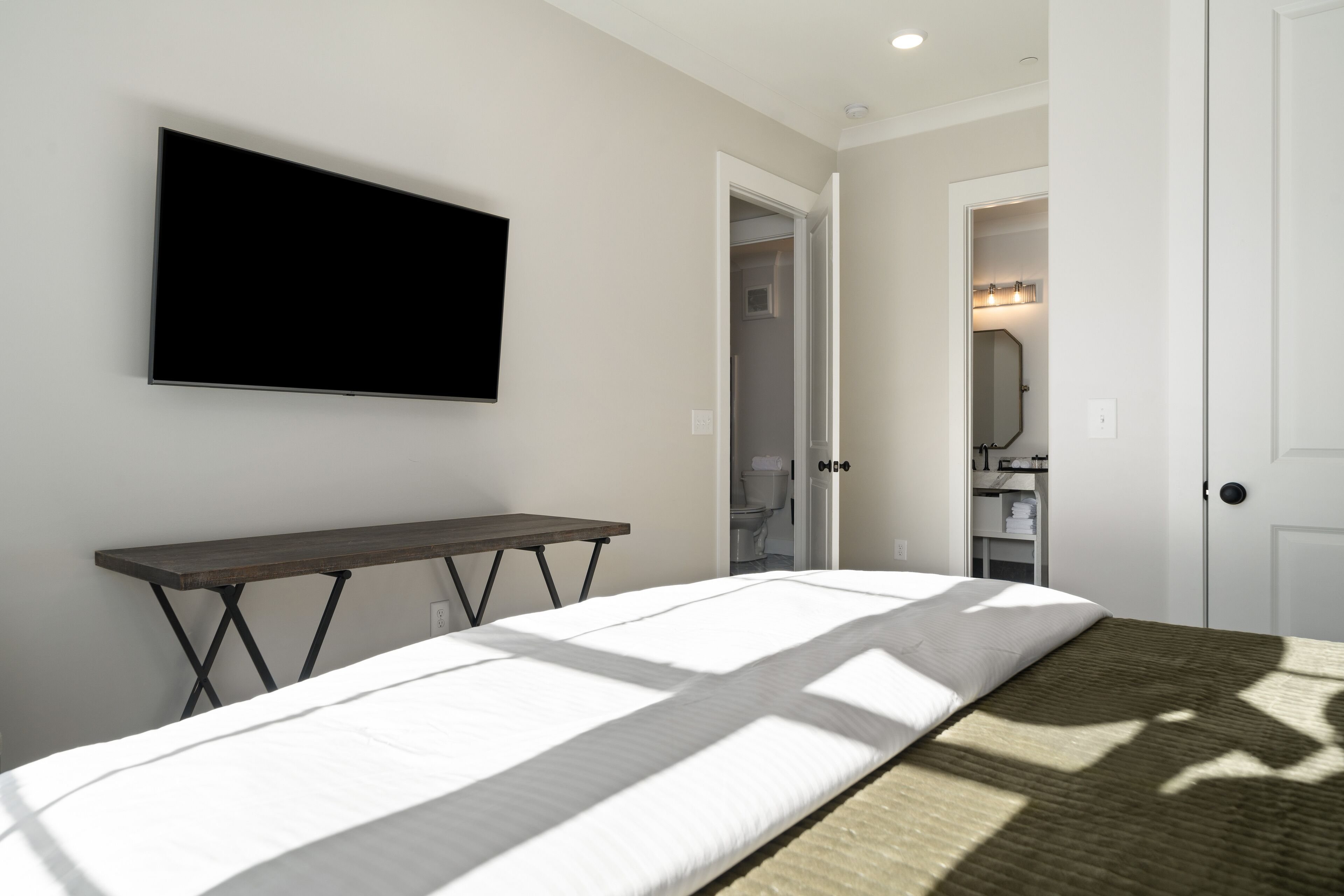 Une chambre à coucher comprenant un lit, un téléviseur fixé au mur, une petite table et une porte donnant sur une salle de bain.