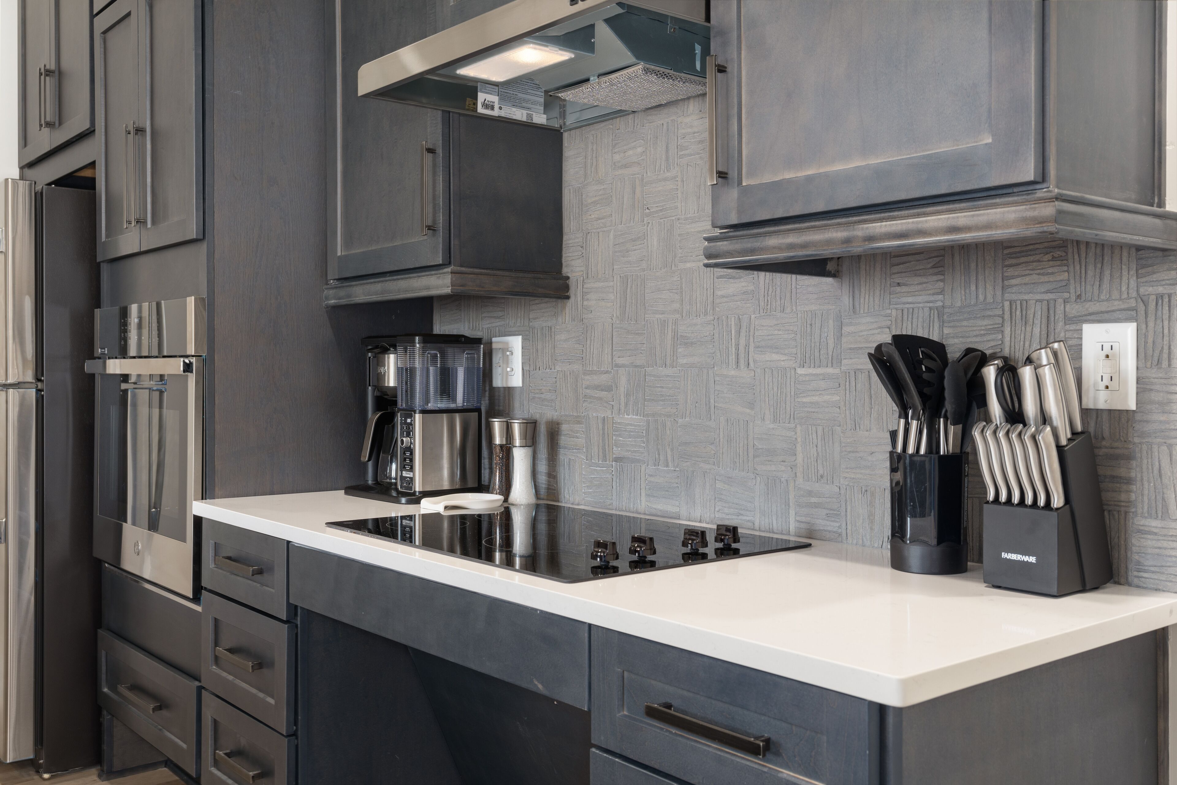 Una cocina moderna con armarios oscuros, encimera blanca, electrodomésticos de acero inoxidable y salpicadero con diseño en espiga.