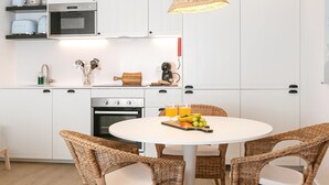 A modern kitchen with white cabinets, a round dining table, wicker chairs, and a hanging light fixture. - Estrelas da Praia - Stylish and Cozy Apartment (Peniche)