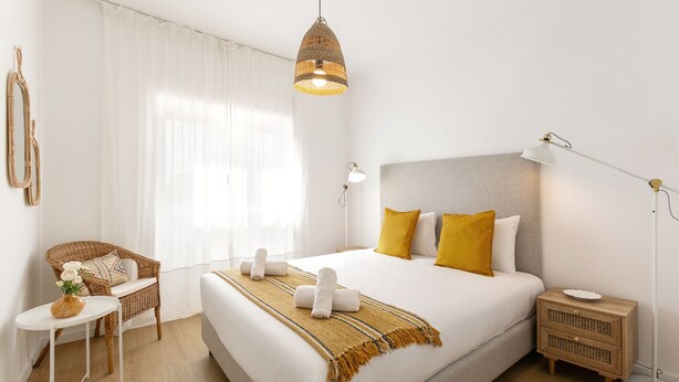 A neatly arranged hotel room with a large bed, a bedside table, a chair, a lamp, and a window with curtains. - Estrelas da Praia - Stylish and Cozy Apartment (Peniche)