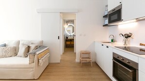 A modern kitchen with white cabinets, a wooden countertop, and a built-in oven. There is a small wooden stool and a white sofa with cushions. - Estrelas da Praia - Stylish and Cozy Apartment (Peniche)