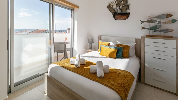 Une chambre d’hôtel moderne dotée d’un grand lit, d’une tête de lit en bois, d’une porte-fenêtre coulissante donnant sur un balcon et offrant une vue sur le paysage urbain.