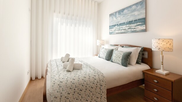 Un lit bien fait, avec des oreillers et une couverture, une table de chevet avec une lampe, et un tableau représentant un paysage marin accroché au mur.