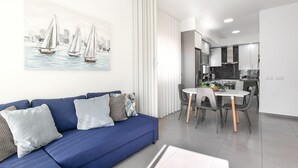 Un salon moderne avec un canapé bleu, un coin repas avec une table ronde et des chaises, et une cuisine en arrière-plan.