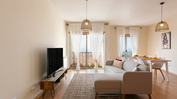 Una sala de estar moderna con televisor de pantalla plana, un sofá, una mesa de comedor con sillas y un balcón con cortinas.