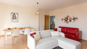 Una sala moderna con un sofá seccional blanco, un sofá rojo, una mesa de comedor con sillas y una cocina al fondo.