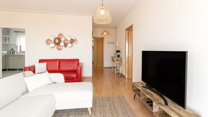 Una sala de estar moderna con un sofá blanco, un sillón rojo y un televisor de pantalla plana grande sobre un soporte de madera. Una lámpara trenzada cuelga del techo, y se observa un plato decorativo en la pared.