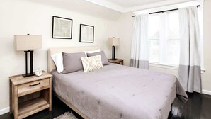 A bedroom with a bed, two bedside tables, a nightstand, a window with curtains, and two wall-mounted picture frames.