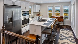 A modern kitchen with white cabinetry, stainless steel appliances, a central island with bar stools, and large windows with a view of trees. - Eagle Ridge - Ridge Road Scenic Cabins - Brimstone Recreation and Big South Fork (Robbins)