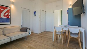 A modern living room with a sofa, a dining table with chairs, a TV mounted on the wall, and a door leading to another room.