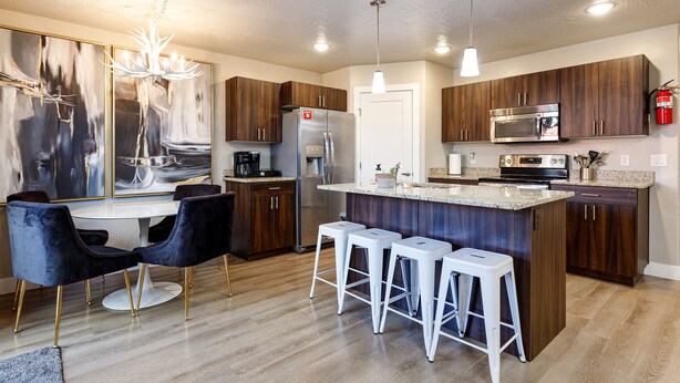 A modern kitchen with a central island, bar stools, dark wooden cabinets, stainless steel appliances, and a chandelier.
