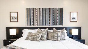 A neatly made bed with patterned pillows, a large striped tapestry, and framed pictures on the walls.