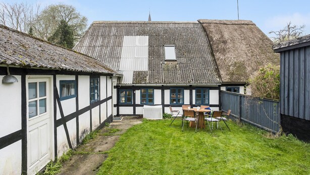 Une maison traditionnelle à colombages avec un toit de chaume, un petit jardin avec une table et des chaises, et un espace clos.
