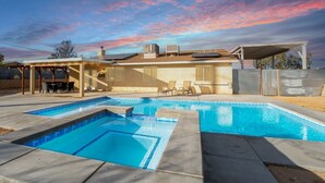 A swimming pool with a pergola and lounge chairs, a covered patio, and a view of the sunset.