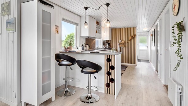 Une cuisine moderne avec des meubles blancs, un îlot central doté de tabourets de bar, un éclairage suspendu et une vue sur l’extérieur grâce à une fenêtre.