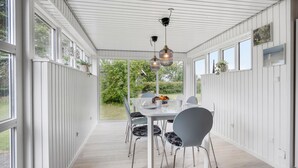 Un espace repas abrité, avec une table et des chaises blanches, de grandes fenêtres et des lampes suspendues.