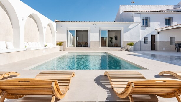 A modern pool area with wooden loungers, a clear pool, and a white building with arches and a tiled roof. - MalagaSuite Villa Nomada El Torcal (Antequera)
