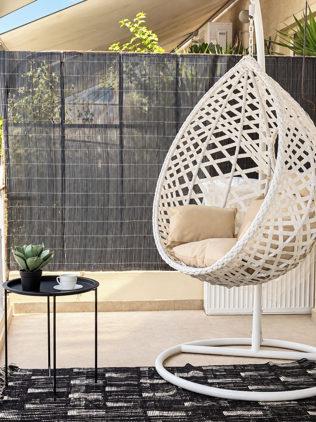 A white woven hanging chair with cushions, a small round table with a cup and a potted plant, and a black woven rug on a balcony with a bamboo fence in the background.