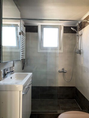 A modern bathroom with a white sink, a mirror, a shower with a handheld showerhead, and two windows. - Funky Boho Kallithea Loft (Kallithea)