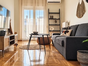 Una sala moderna con un sofá gris, piso de madera, un televisor y un balcón con cortinas.