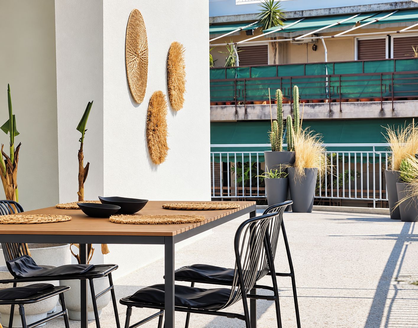 Ein Außenbereich zum Essen mit einem Holztisch, schwarzen Stühlen und gewebten Tischsets. Im Hintergrund sind Kübelpflanzen und ein Geländer zu sehen.
