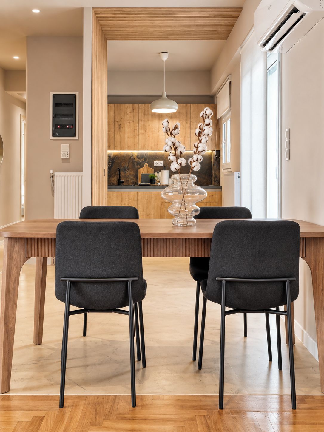 A modern dining area with a wooden table and four chairs, a vase with flowers, and a kitchen in the background.
