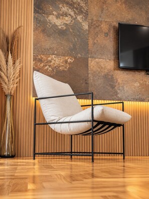 Una silla moderna con estructura metálica y cojines blancos, ubicada frente a una pared de madera con listones verticales y un detalle de pared de piedra texturizada. Un televisor de pantalla plana está montado en la pared sobre la silla.