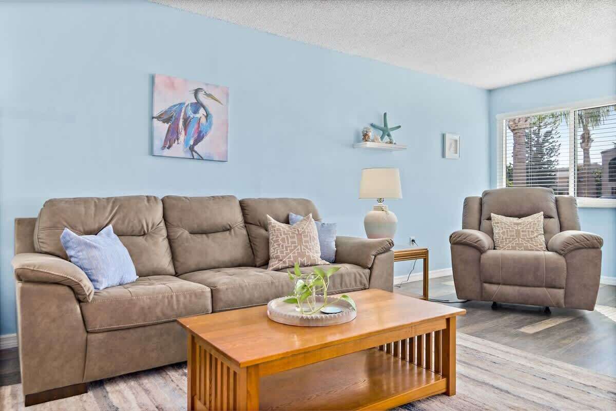 A living room with a beige sofa, a wooden coffee table, and a beige armchair. There is a painting of a bird on the wall, a starfish decoration, and a lamp on a side table.