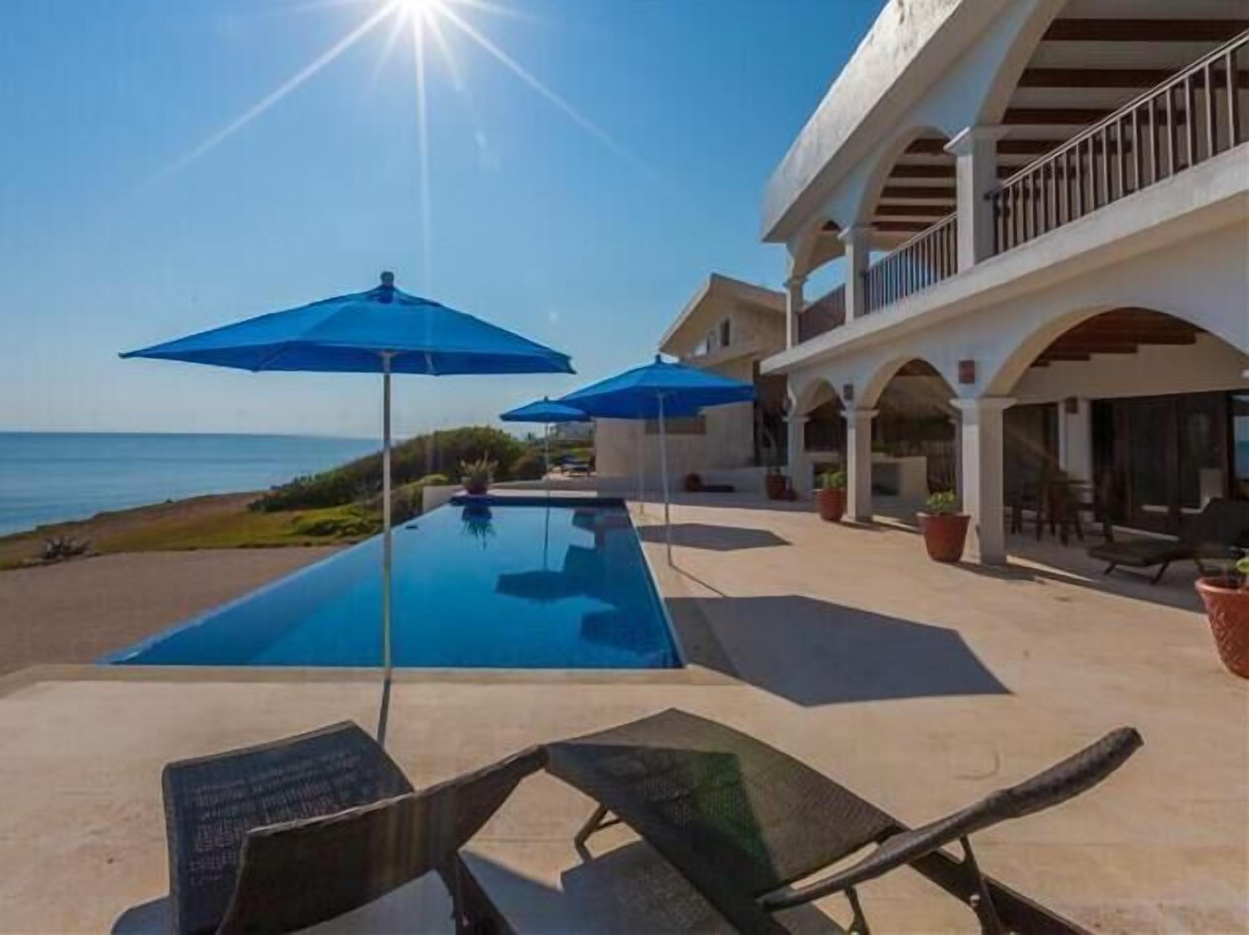 A swimming pool with blue umbrellas, lounge chairs, and a view of the sea.