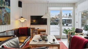A cozy living room with a sofa, coffee table, and a television. There is a bookshelf and a window with a view of a snowy landscape. - 4 bedroom gorgeous home in Hornbæk (Hornbæk)