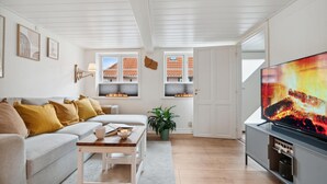 Un salon moderne avec un canapé, une table basse et un téléviseur. Il y a des fenêtres donnant sur des immeubles et une plante est posée sur le sol.