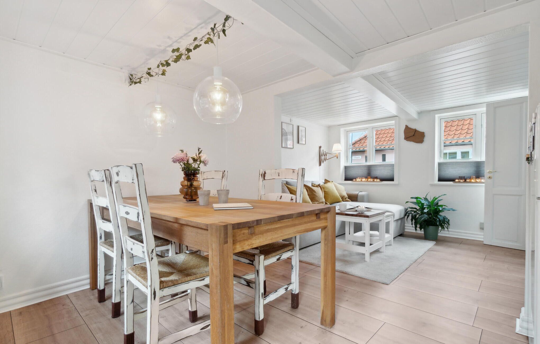 A dining area with a wooden table and white-painted chairs, a vase with flowers, and hanging pendant lights.