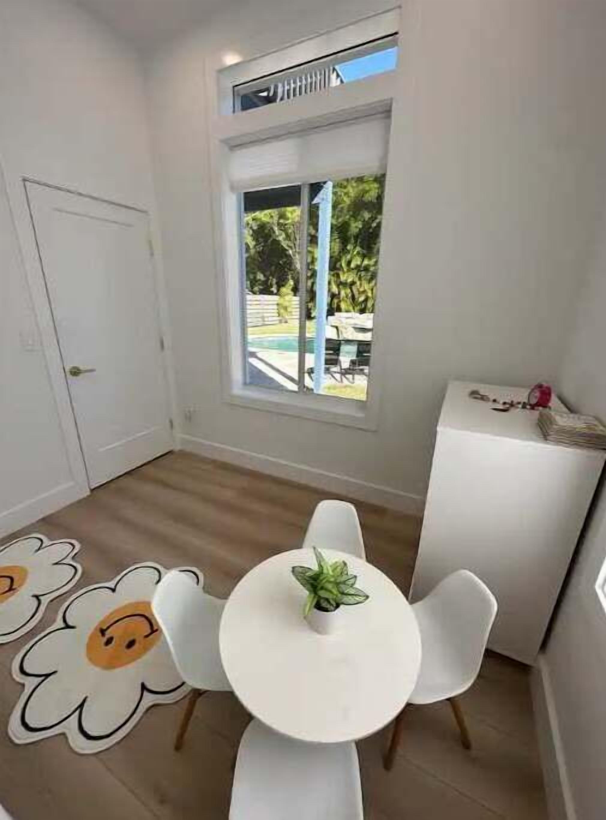 A small, bright room with a white table and chairs, a window with a view of a pool and greenery, and a white door.