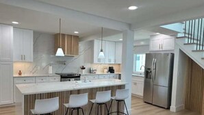 A modern kitchen with white cabinets, a marble countertop, and a stainless steel refrigerator. There are three bar stools and a large island with a sink. - Private Resort (Sarasota)