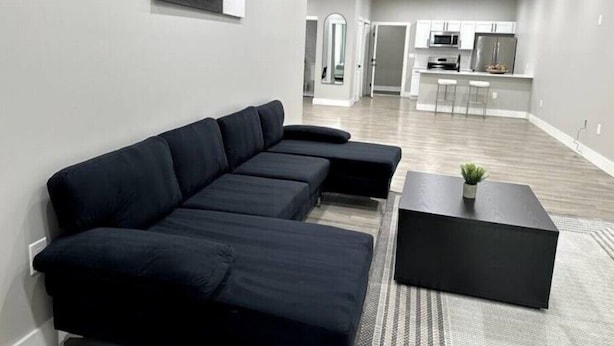 Un salon moderne avec un canapé d’angle noir, une petite table basse noire et un coin cuisine en arrière-plan.
