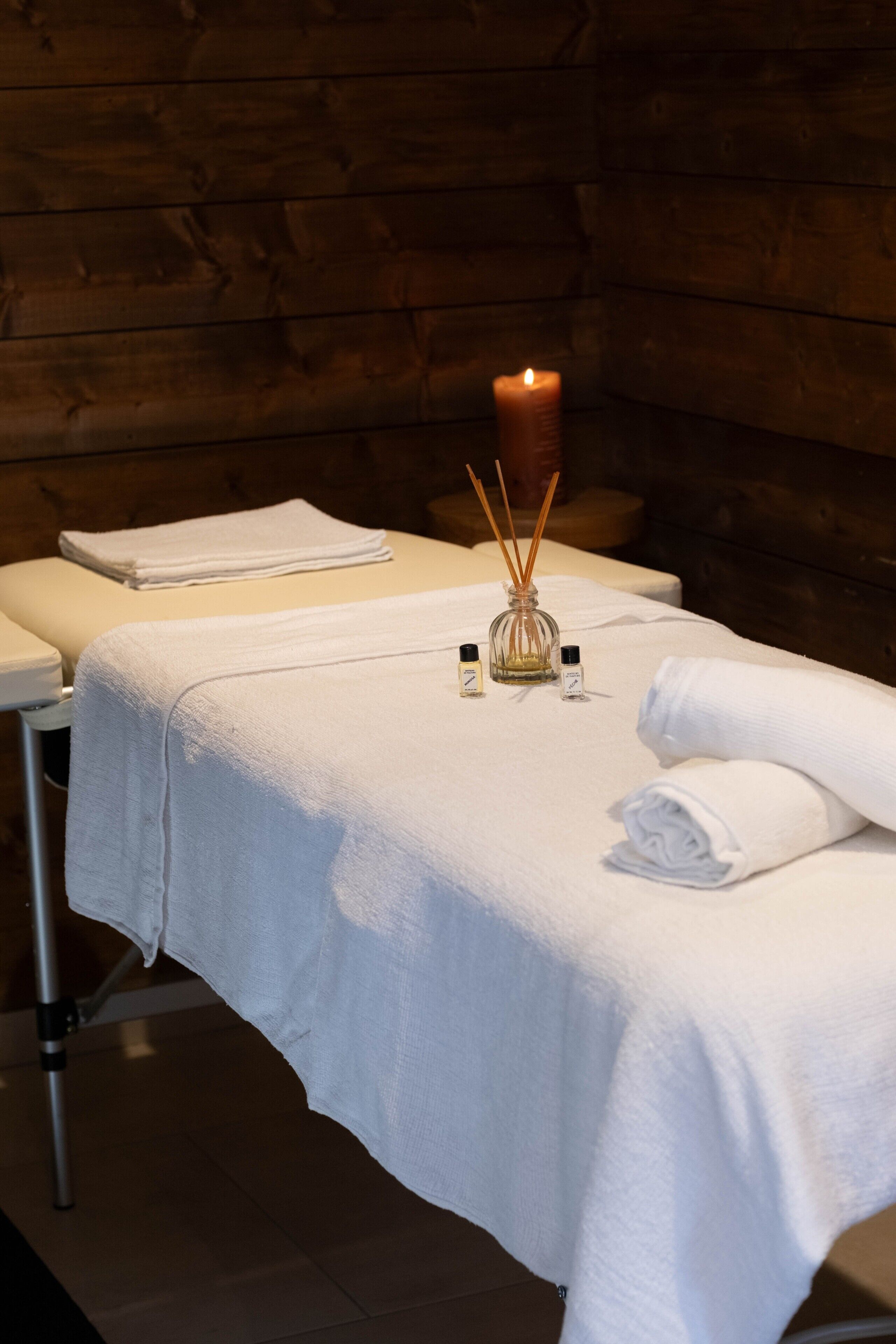 Une table de massage recouverte d’une serviette blanche, une bougie allumée et deux petits flacons d’huiles essentielles.