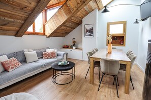 A loft-style room with a wooden ceiling, a sofa, a dining table with chairs, a small round table, and a TV mounted on the wall.
