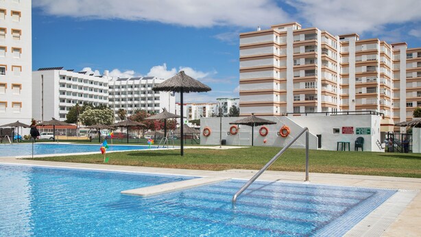 A swimming pool with a lifeguard stand, a thatched umbrella, and a multi-story building in the background.