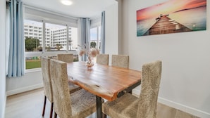A dining area with a wooden table and woven chairs, a painting of a pier, and a view of buildings through large windows.