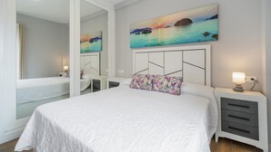A bedroom with a bed, a nightstand, a mirror, and a painting of a sunset over water.