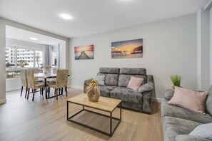A modern living room with a grey sofa, a wooden coffee table, and a dining area with wicker chairs.