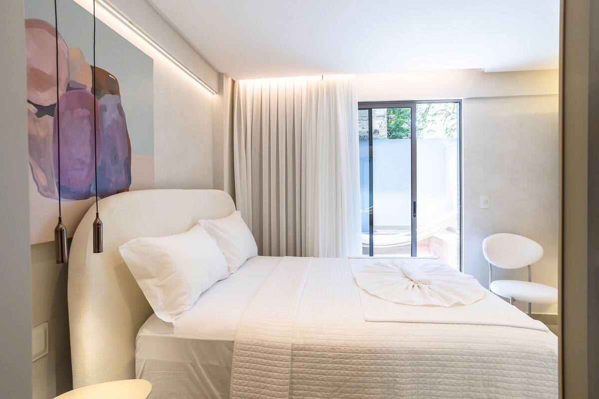 A modern hotel room with a large bed, a chair, a painting on the wall, and a view of the outdoors.