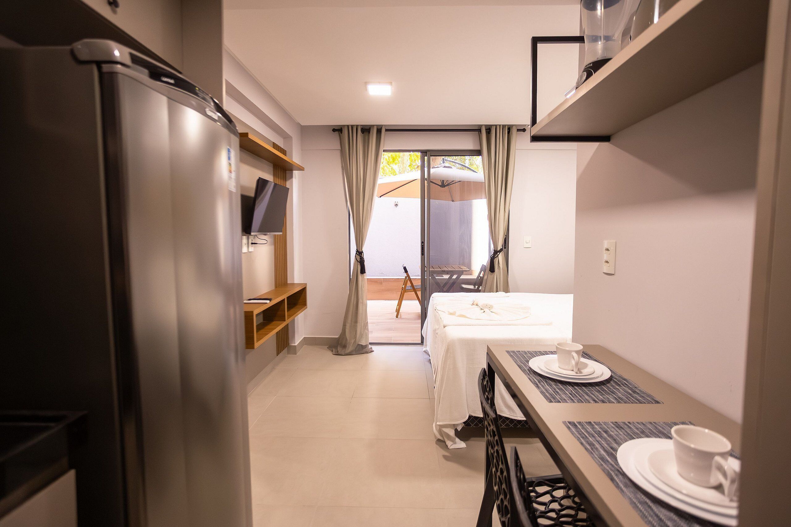 A compact hotel room with a kitchenette, a bed, a dining table with a cup and saucer, and a sliding glass door leading to an outdoor area.