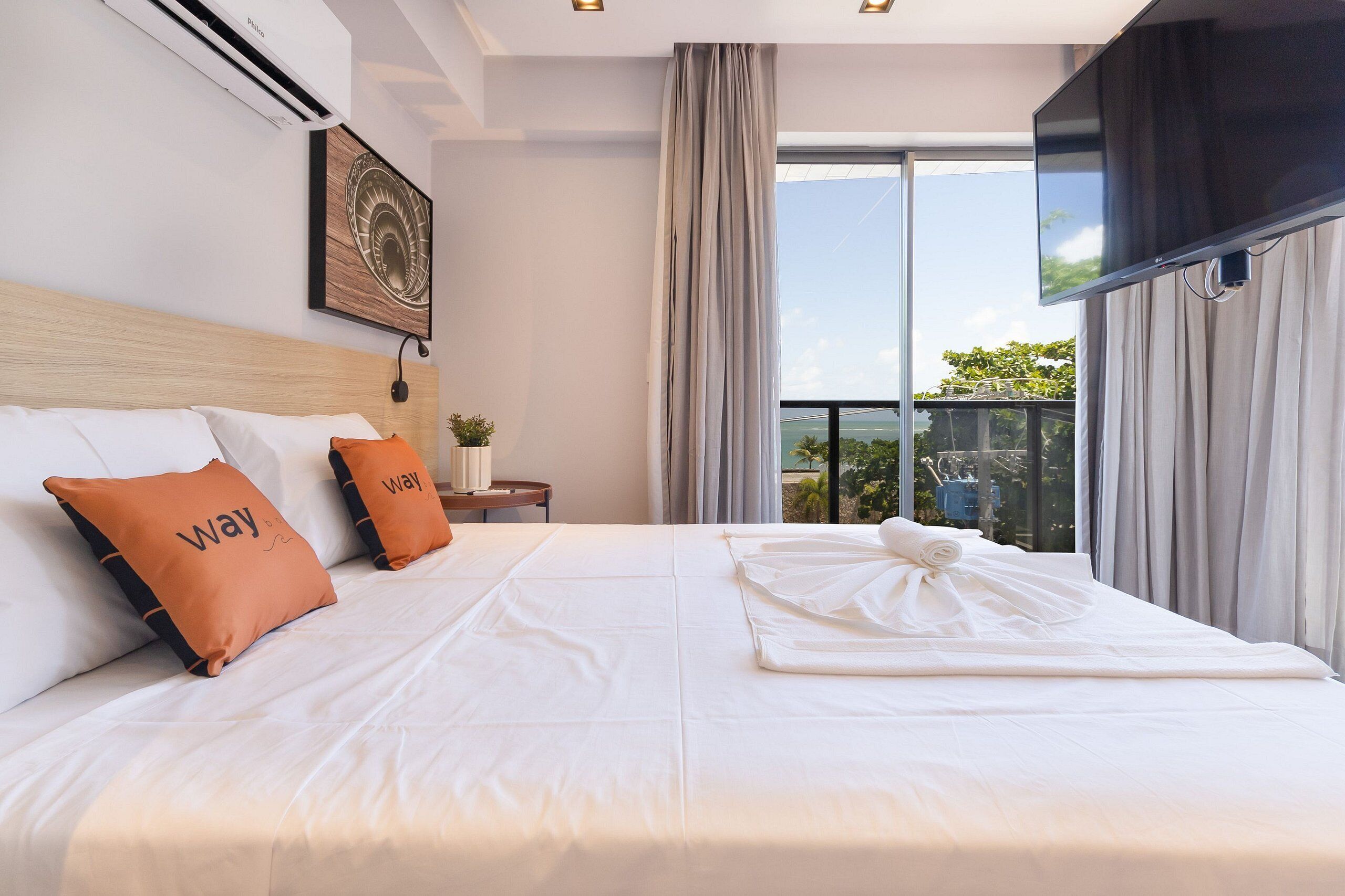 A hotel room with a large bed, two pillows with the word 'Way' on them, a flat-screen TV mounted on the wall, and a window with curtains.