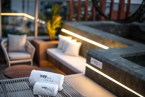 A well-lit outdoor seating area with a sofa and chairs, a small table with towels, and a stone wall.