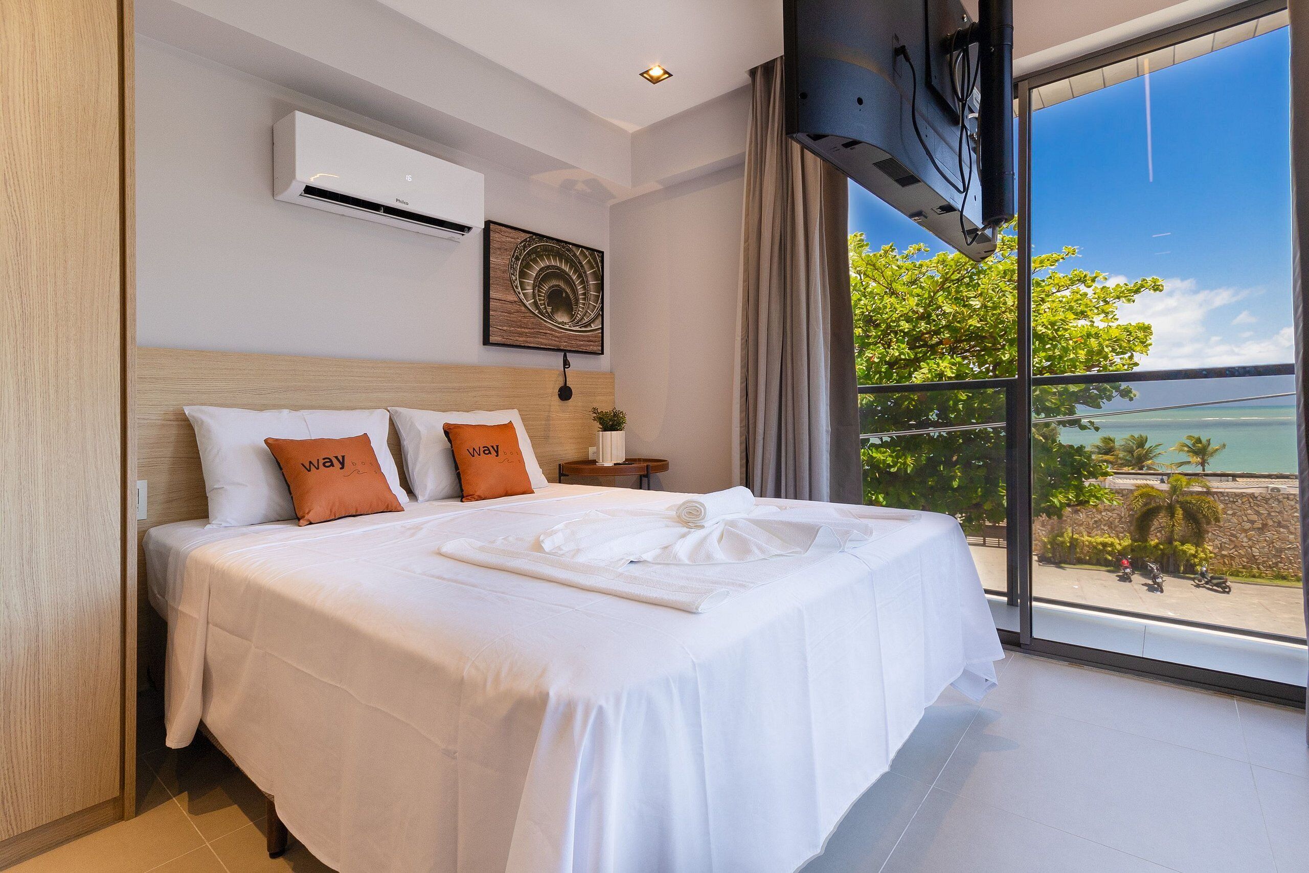 A hotel room with a large bed, an air conditioning unit, a view of the outdoors, and a window with curtains.