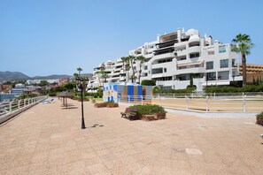 A coastal promenade with modern buildings, a palm tree, and a clear sky.