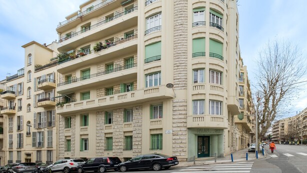 A multi-story building with balconies, a ground-floor entrance, and a crosswalk.