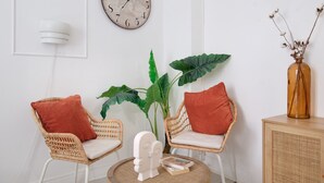 A room with two wicker chairs, a large plant, a clock, and a vase with dried flowers.